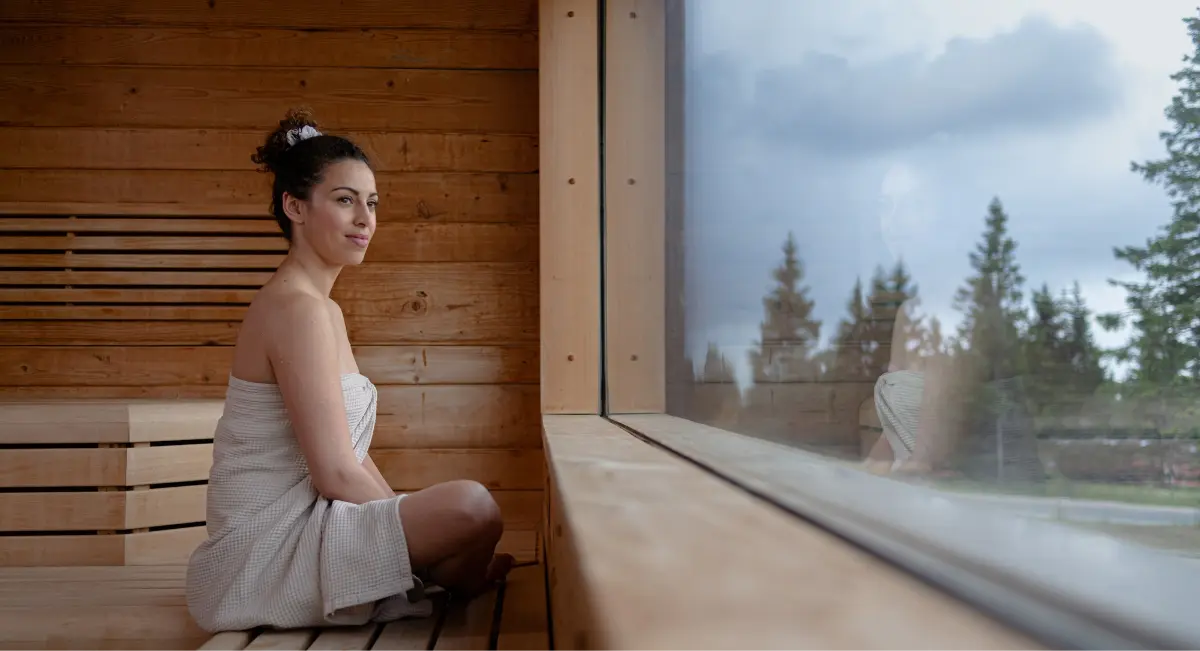 Ženska, zavita v brisačo, sedi v leseni savni ob oknu.