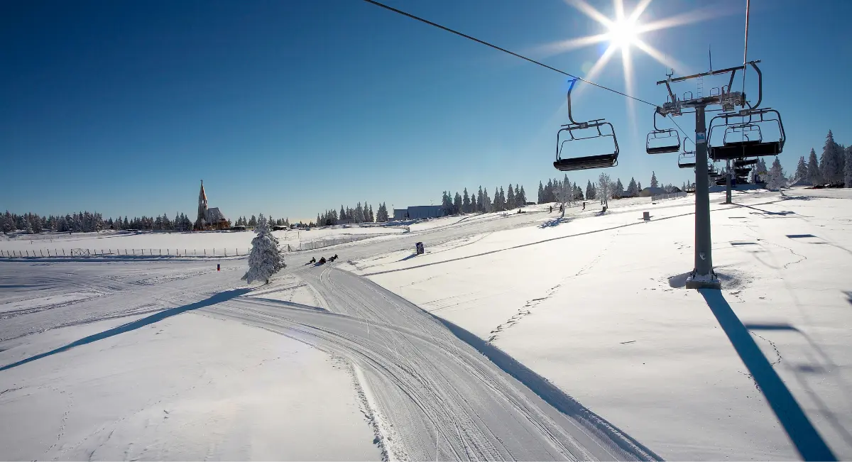 Pogled na snežno pokrajino s sončnim dnevom, smučarsko progo in žičnico.