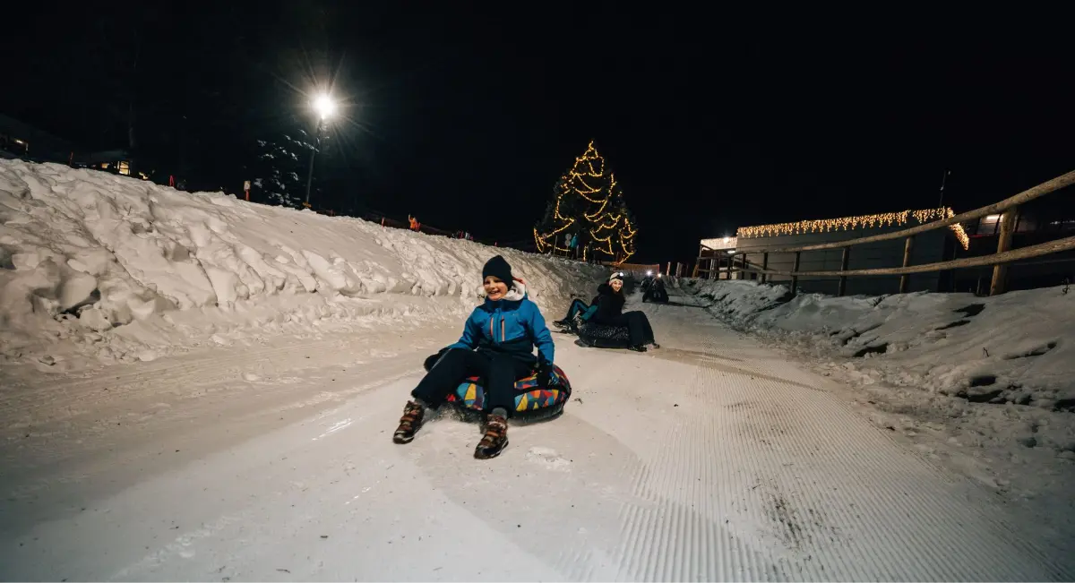 Otroci se sankajo ponoči na zasneženem hribu, obdan z lučkami in božičnim drevesom.