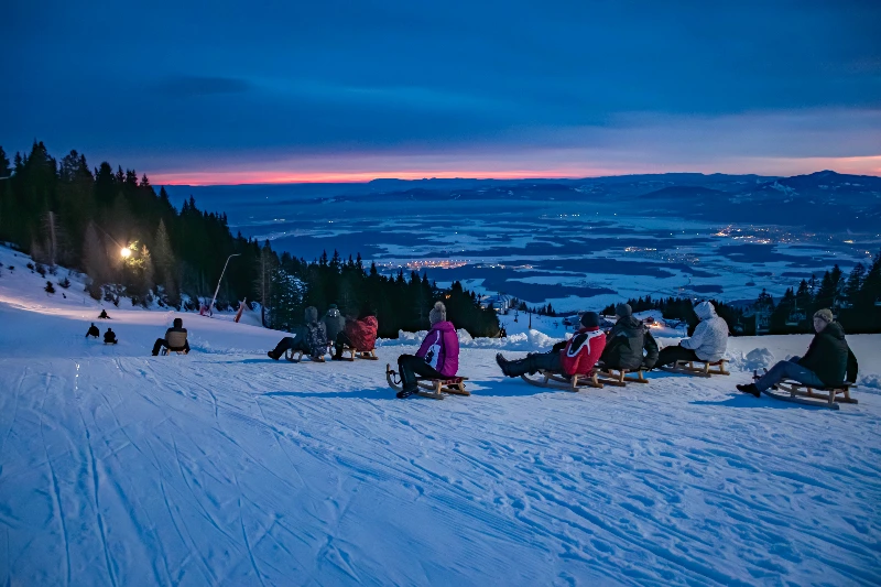 Skupina ljudi na saneh na zasneženem hribu ob zori z razgledom na dolino.