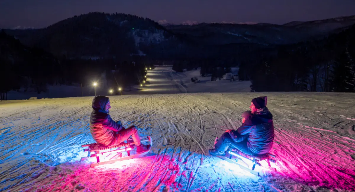 Dve osebi sankata ponoči na osvetljenem smučišču, z barvno osvetlitvijo sank.