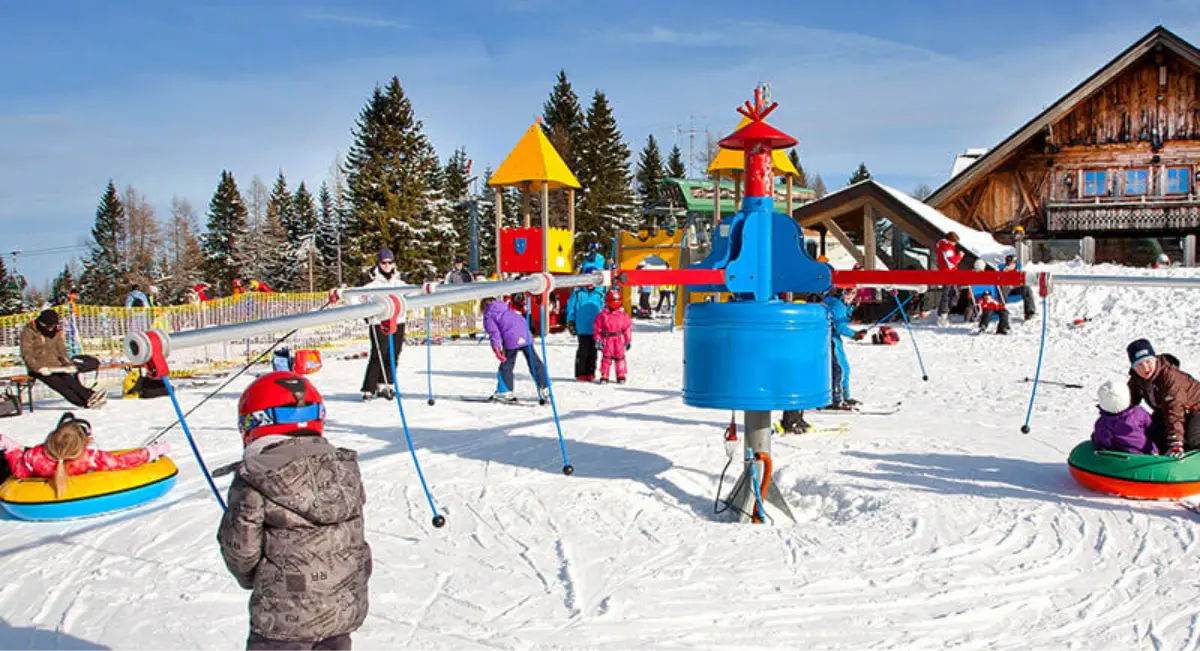 Otroci se igrajo na snežnem igrišču z igrali in hiškami v ozadju, obdani z zasneženo pokrajino in leseno kočo.