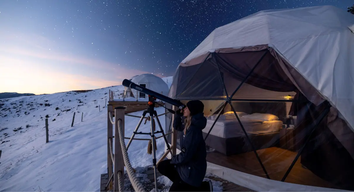 Oseba z daljnogledom pred kupolastim šotorom na zasneženi pokrajini, pod zvezdnatim nebom.